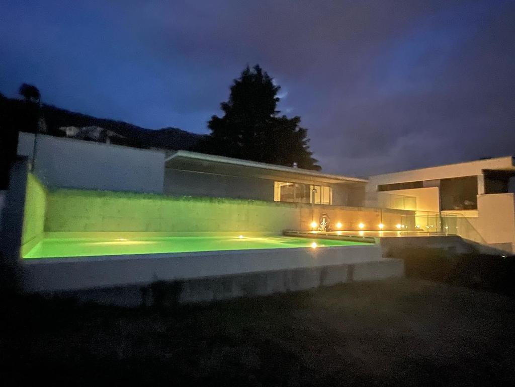 Modern two-story house with a swimming pool illuminated by colorful lights at night. The house has a minimalist design with large windows and a sleek, contemporary appearance.