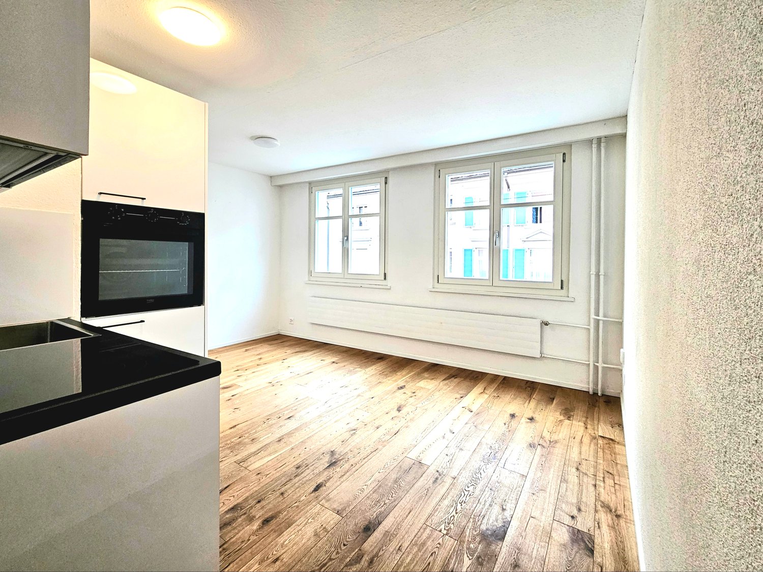 Empty room with wooden floor, white walls, two windows, built-in oven, and a sink.