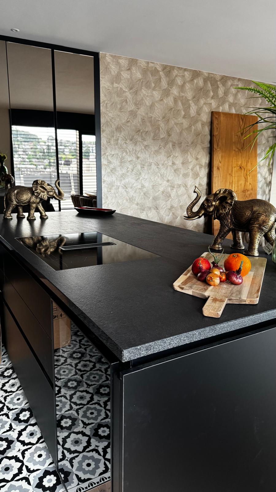 modern kitchen with black granite counter top, white cabinets, patterned floor, large window with blinds, elephant sculptures, cutting board with fruits and vegetables