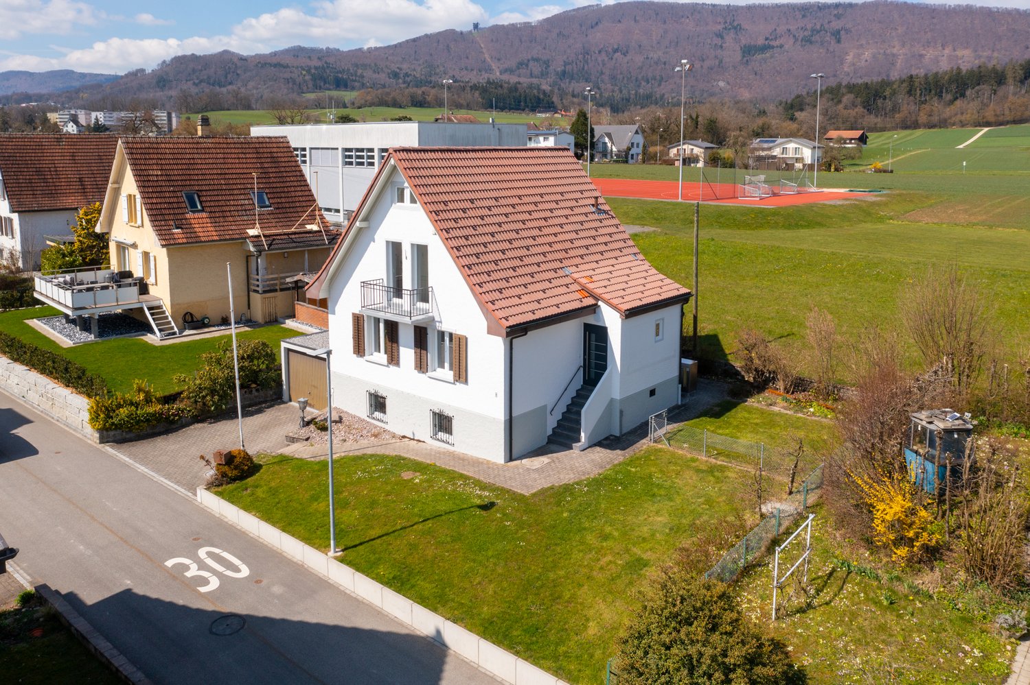 Detached house, brown roof, white walls, balcony, garage, garden, tennis court in the background
