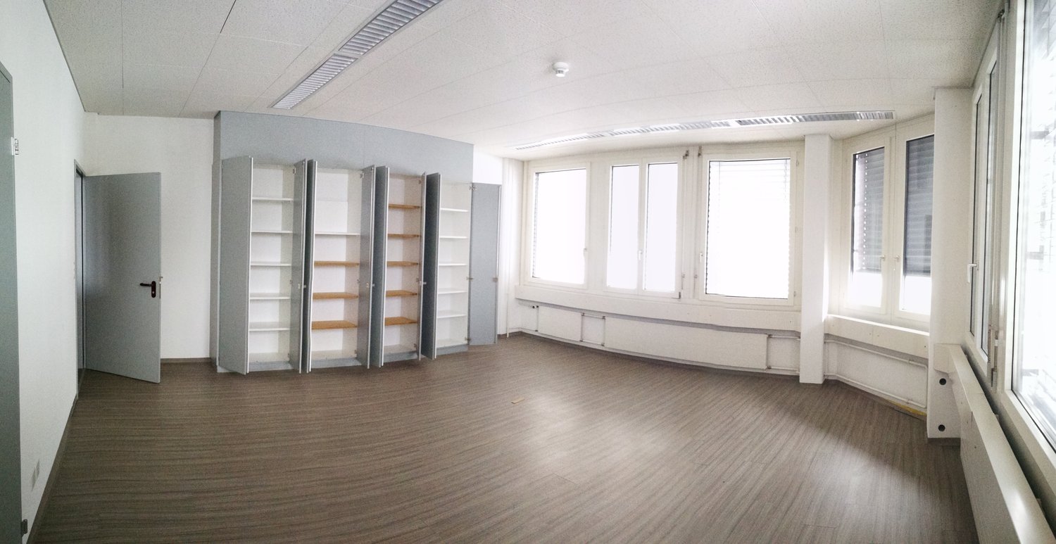 Large open room with white walls, hardwood flooring, and multiple built-in shelving units along the walls. The room has several large windows providing natural light.