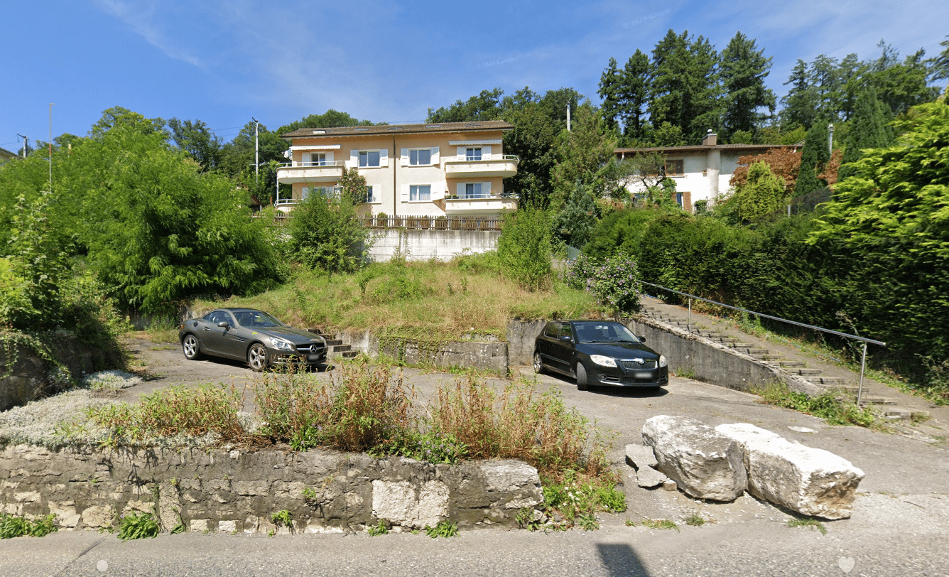 2-story apartment building with balconies, surrounded by trees and greenery, with a parking area in the foreground containing two cars