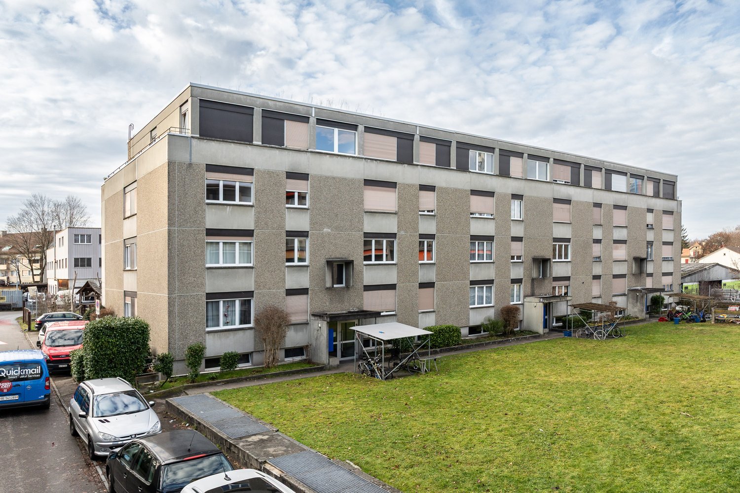 4 story apartment building, grey exterior, multiple windows, bicycles parked outside, green lawn in front