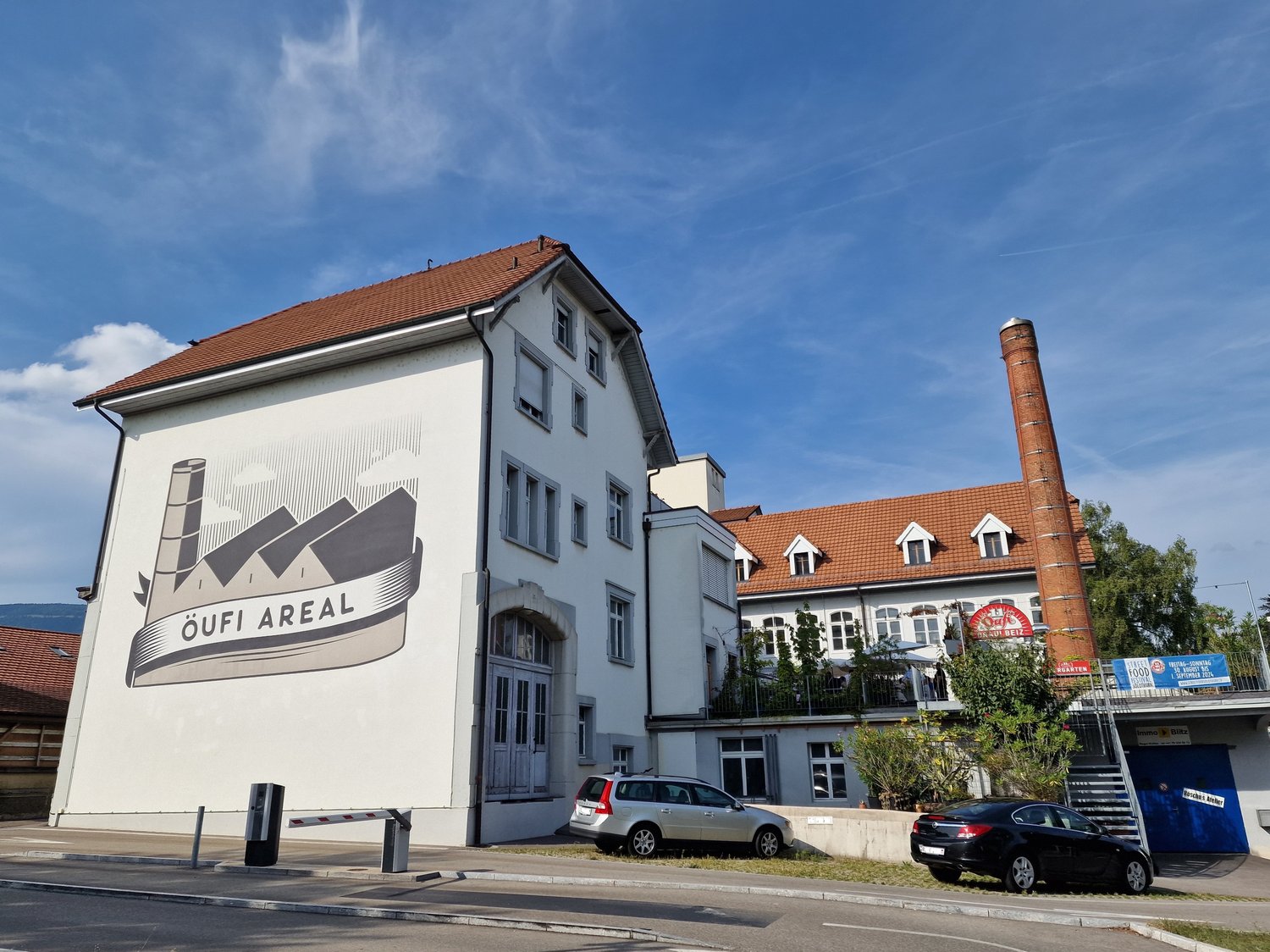 A multi-story building with a white exterior and a red tile roof. The building has several windows and a large sign on the side that says 'OUFI AREAL'. There are some cars parked in front of the building, and a tall chimney or smokestack can be seen in th