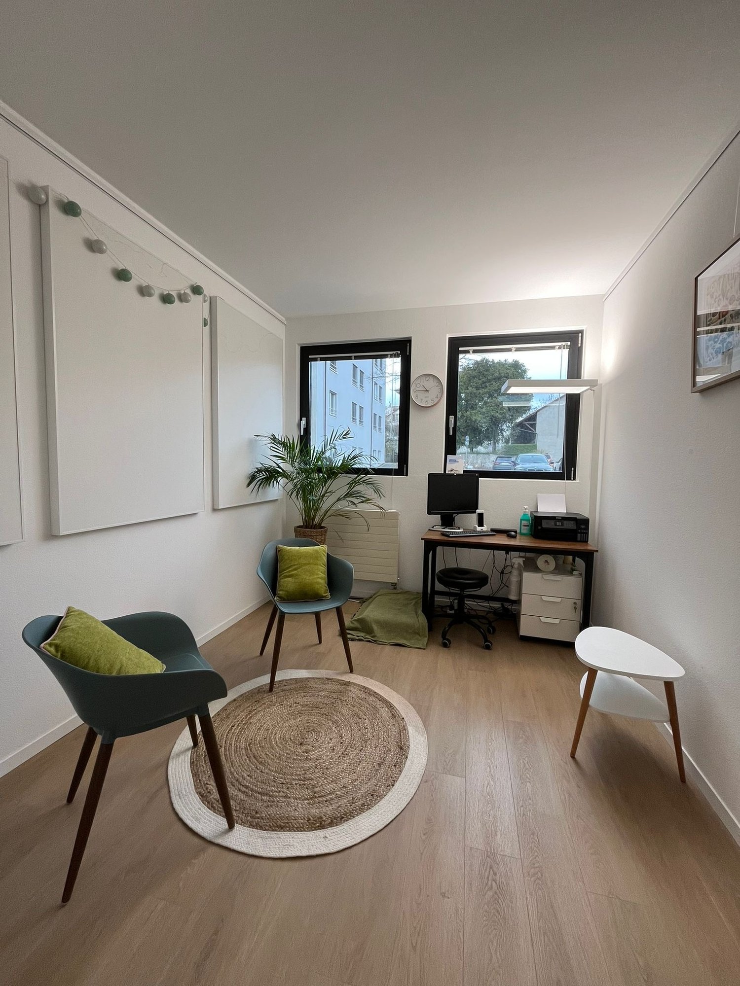 small room with wooden floor, white walls, two chairs, round rug, desk with computer, radiator, plant, clock, picture frame