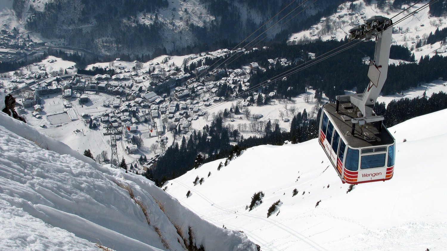 Aerial view of a village on the mountainside covered in snow with houses, trees, and a cable car going through it.