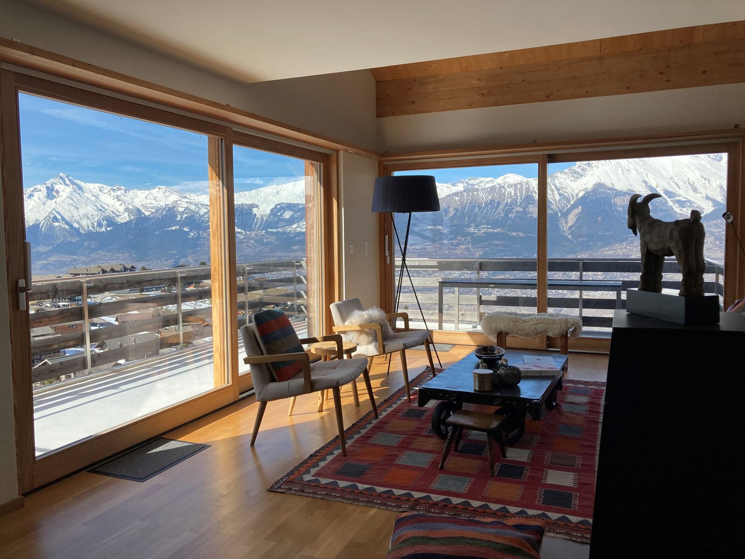 large glass windows, wooden floor, carpet, glass door to balcony, coffee table, chairs, statue, rug, mountains view