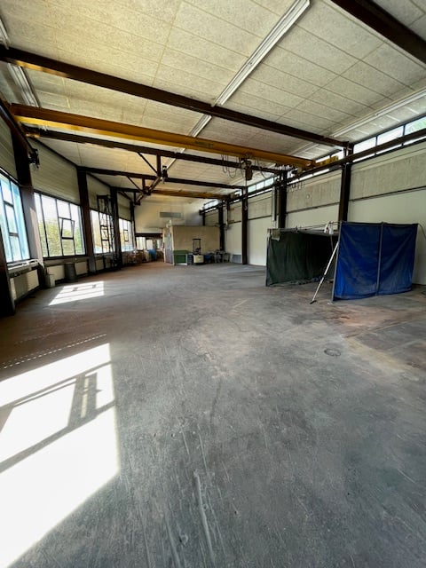 Large open space with concrete floor, exposed ceiling beams, and windows along the walls. The space appears to be a warehouse or industrial facility with some equipment and storage items visible.