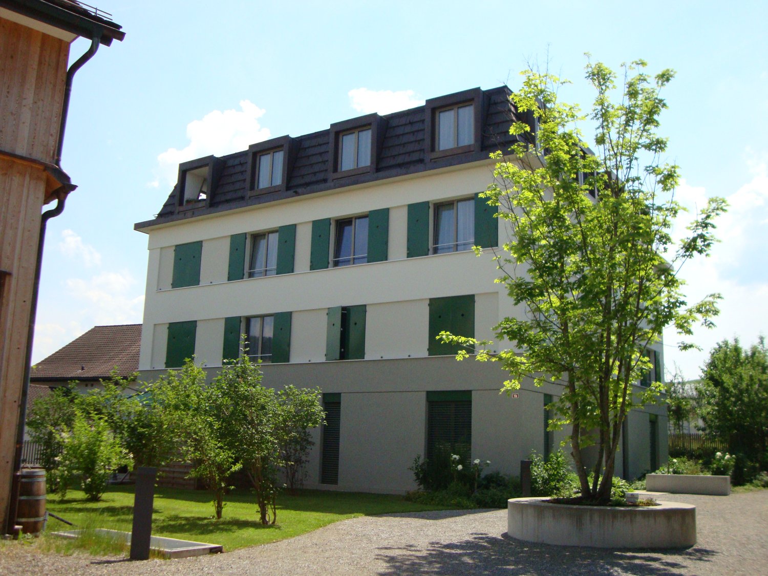 3 story apartment building, white walls, green shutters, gravel driveway, tree, plants, barrel