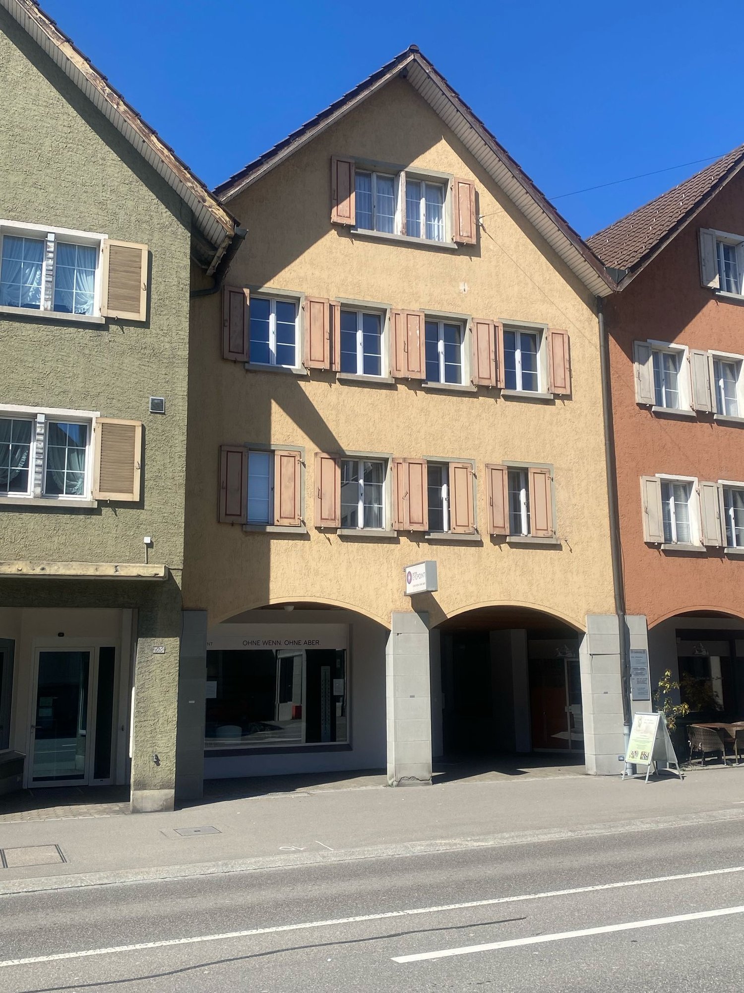 The image shows a multi-story building with a traditional European architectural style. The building has a yellow exterior with brown shutters on the windows. The roof has a gabled design with brown tiles. The ground floor has a commercial space with a si