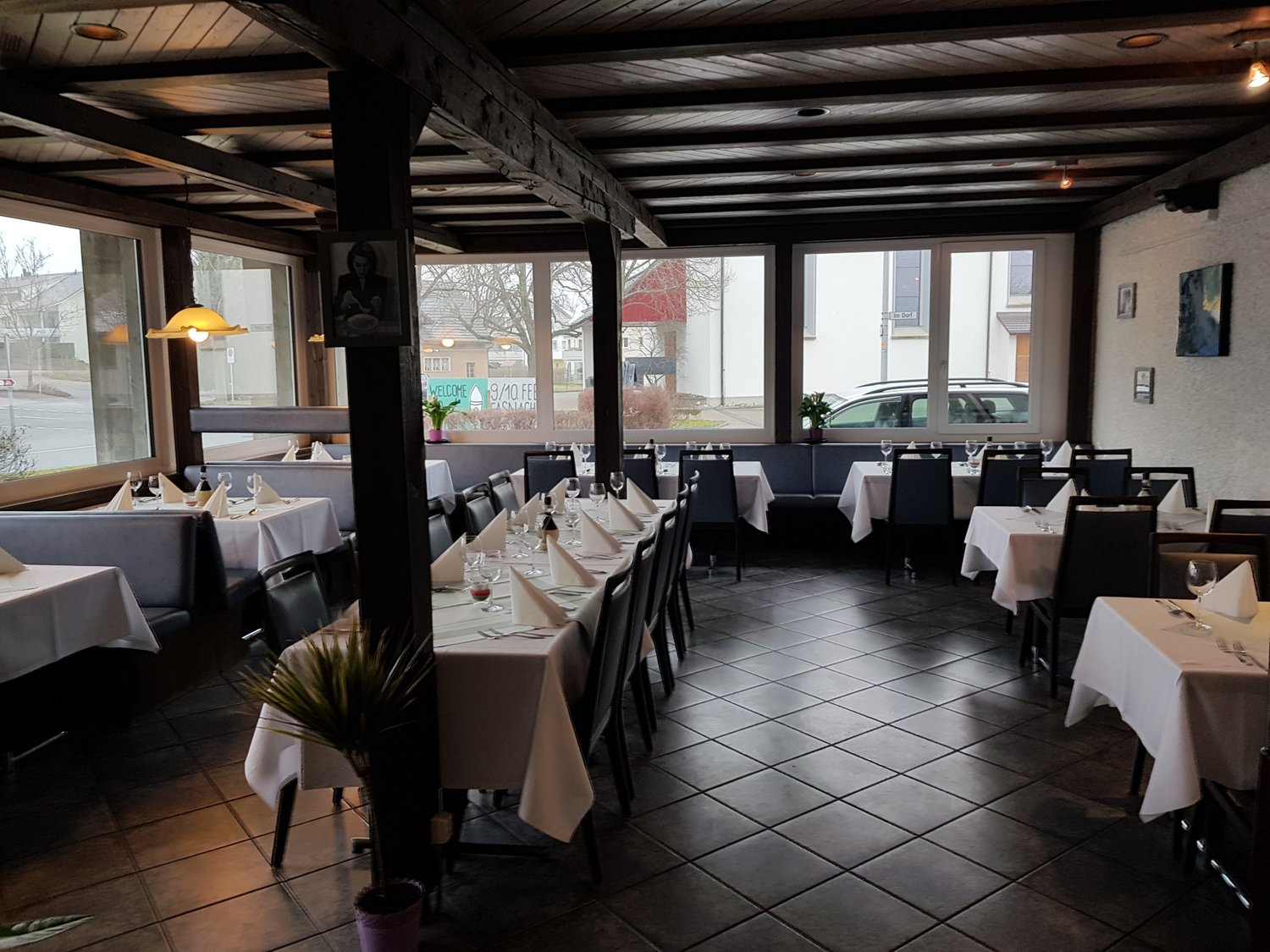 A restaurant interior with wooden beams, large windows, dark tiled floors, and tables with white tablecloths and neatly arranged wine glasses and napkins.