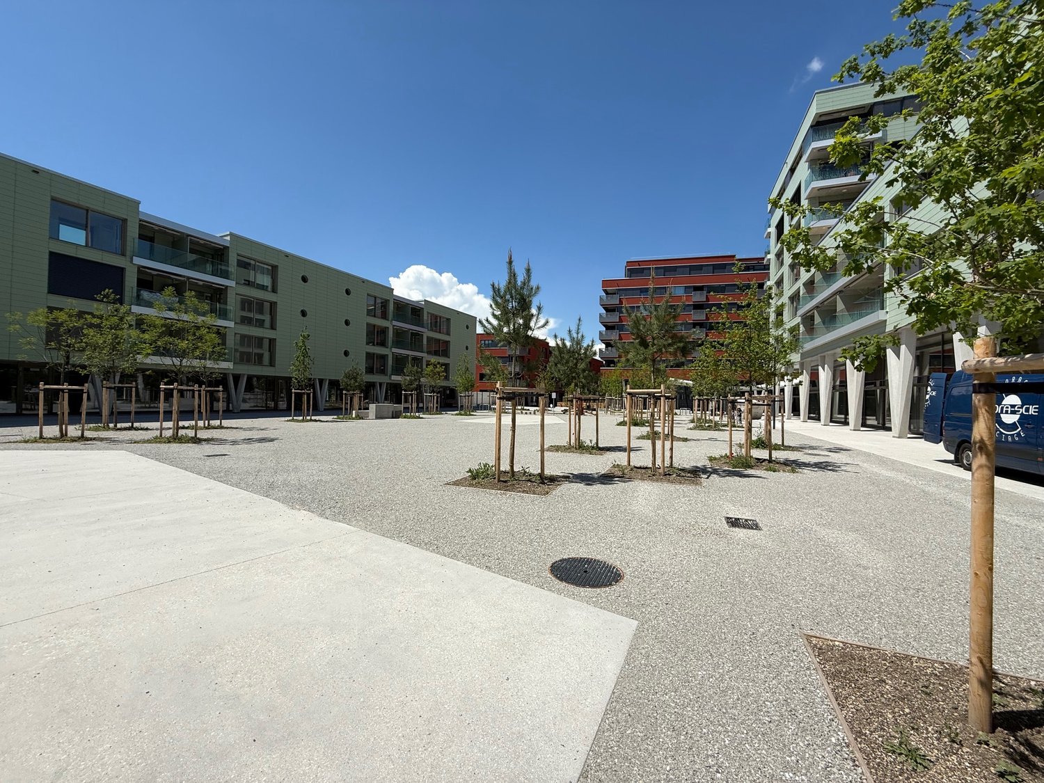 The image shows a modern residential area with multi-story apartment buildings. The buildings have balconies and are surrounded by a paved plaza with trees and benches. The area appears to be well-designed and maintained, with a clean and open layout.