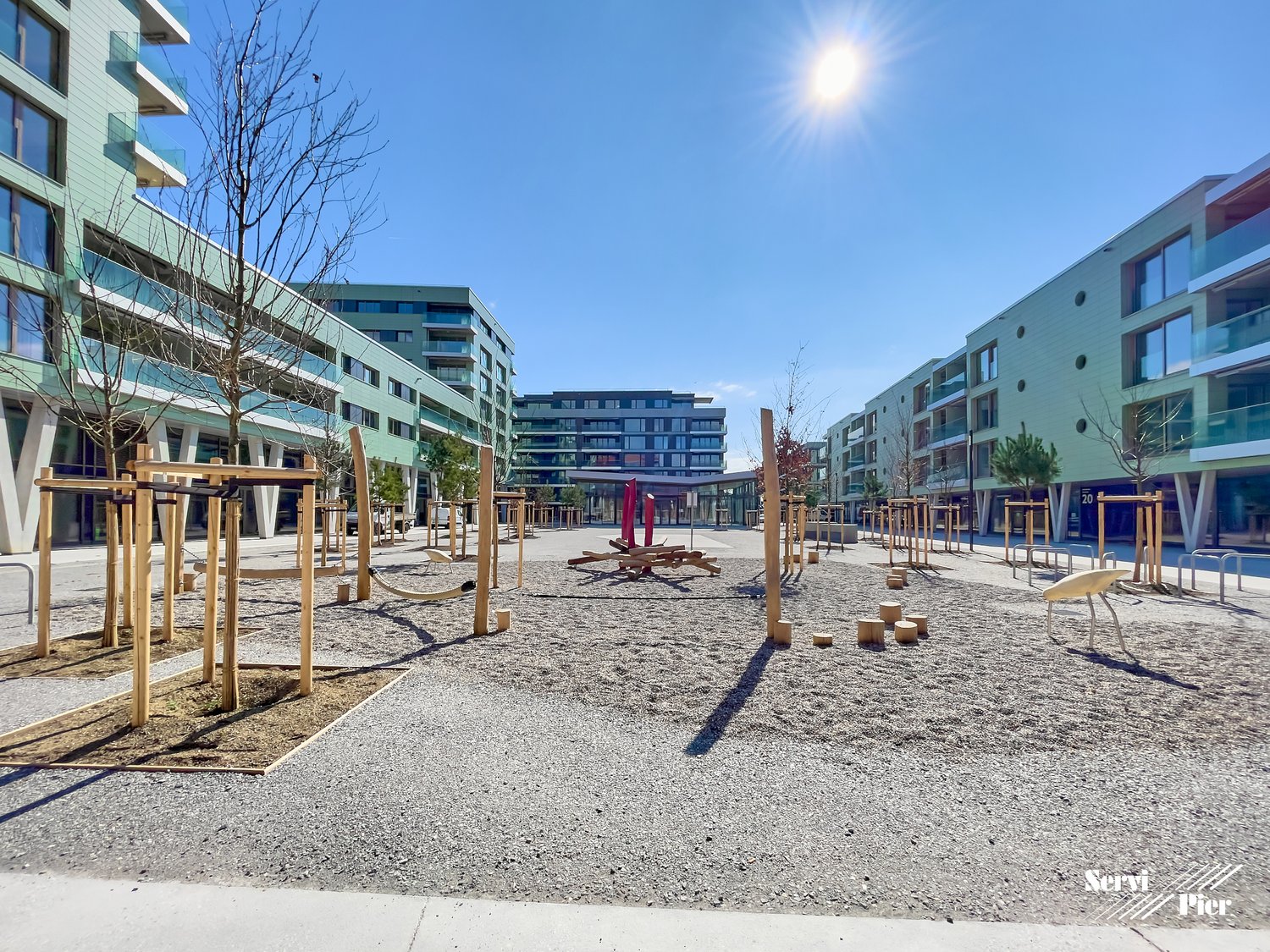 The image shows a public plaza or outdoor space surrounded by modern multi-story residential buildings. The plaza features wooden benches, sculptures, and a gravel-covered ground. The buildings have balconies visible, indicating they are likely apartments