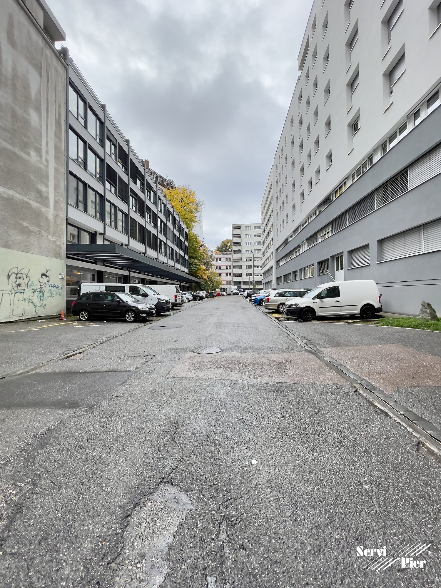 A street with parked cars along the sides, two tall buildings, concrete sidewalk, a few trees, and cloudy sky