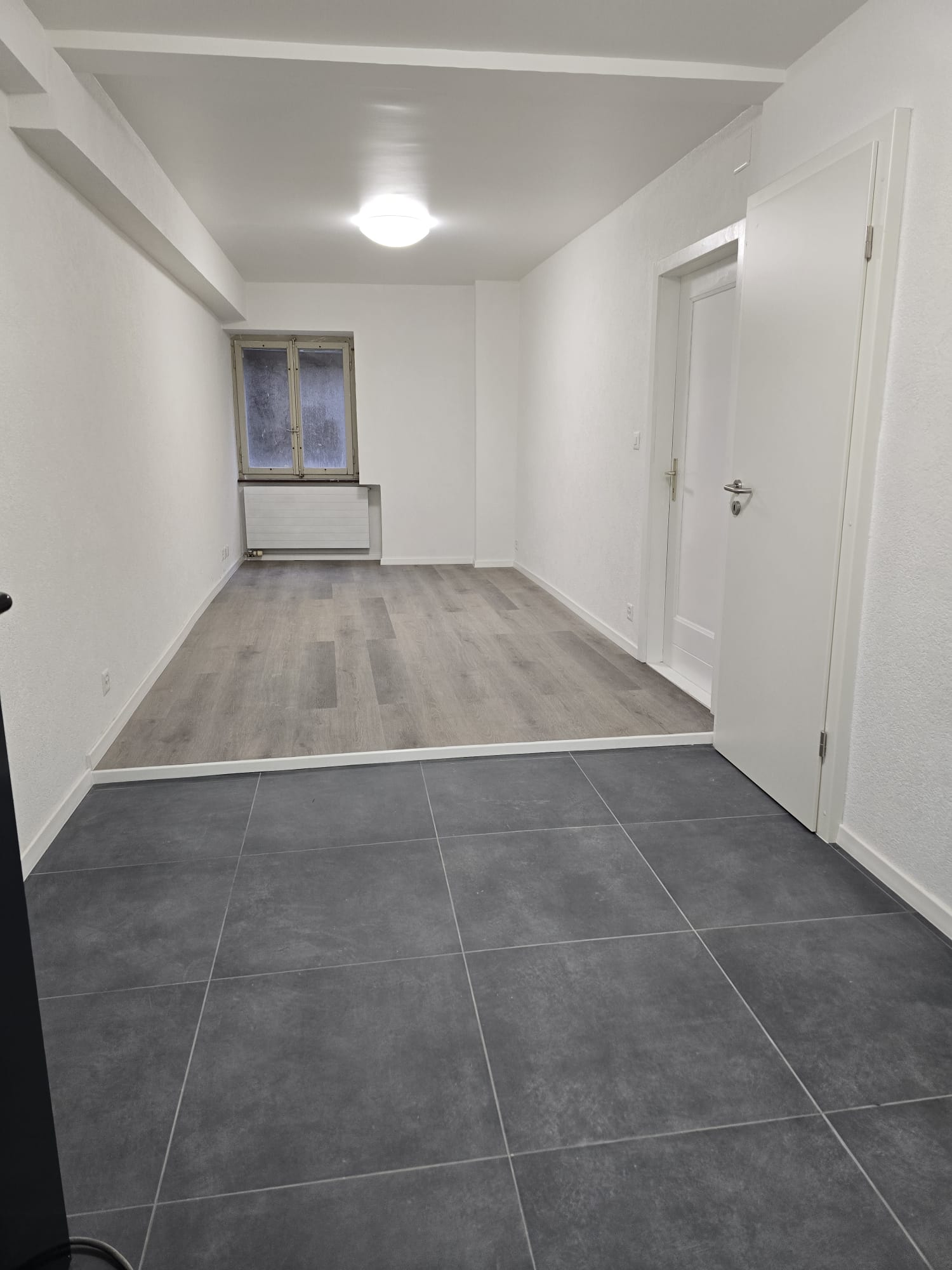 An empty room with tiled flooring, white walls, and a single window. It features a ceiling-mounted light and an open door. There is a radiator beneath the window and two power outlets on the wall.