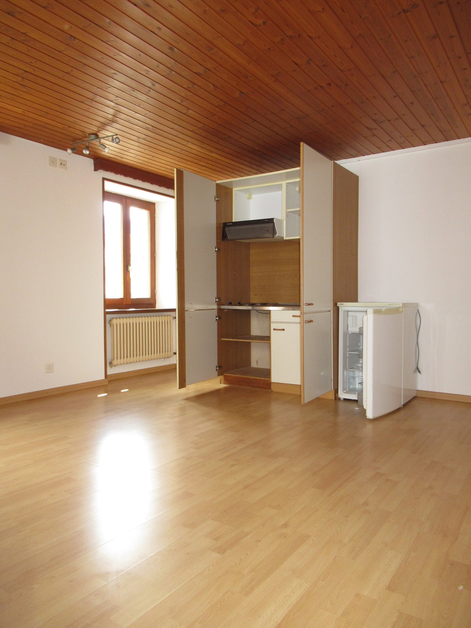Empty room with wooden floor, white walls, kitchen area with open cabinets, window, and a refrigerator.