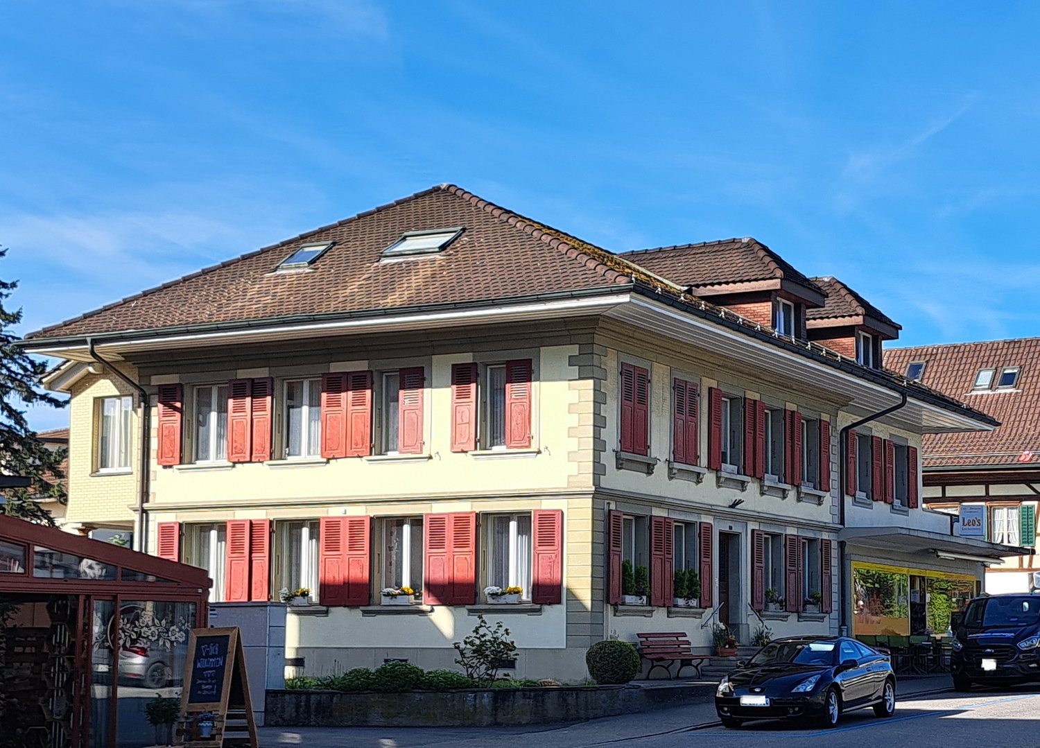 Building has brown tiled roof, yellow walls, and red shutters on windows, garden area with plants, bench, and a car parked nearby