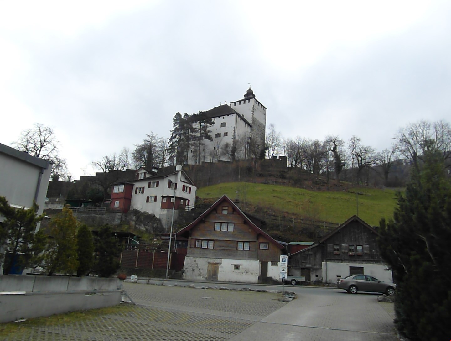 Several houses on hill, larger one at top, brown wooden houses below, small town with cars