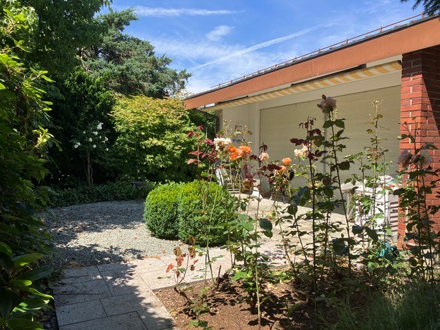 Garden with gravel path, various plants and flowers, brick building with garage, and a white chair.