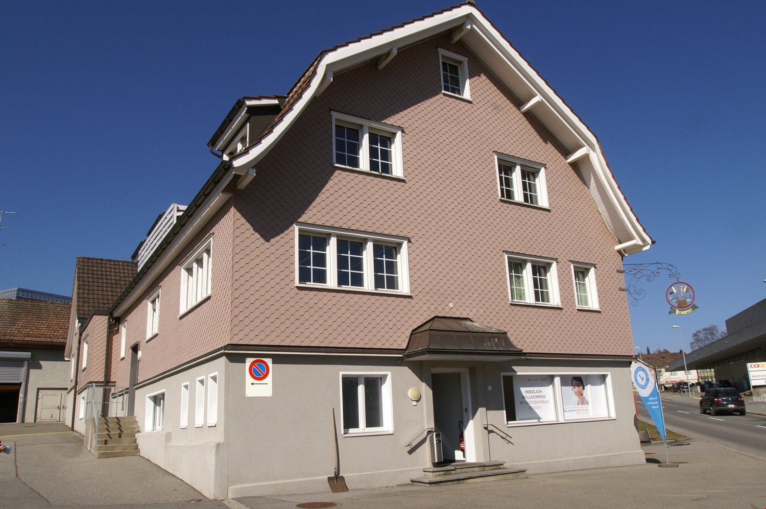 3-story building with a brown tiled roof, white trim, and multiple windows. The building has a commercial space on the ground floor and appears to be located in a town or city setting.