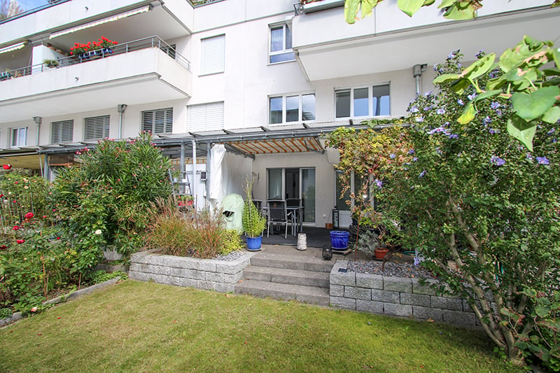 A white building with balconies, a garden in the front, a covered seating area with a table and chairs.