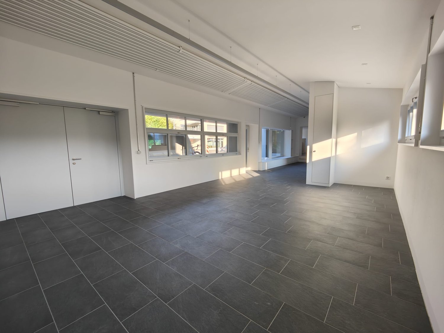 Empty room with white walls, gray tiled floor, large windows, and two white doors