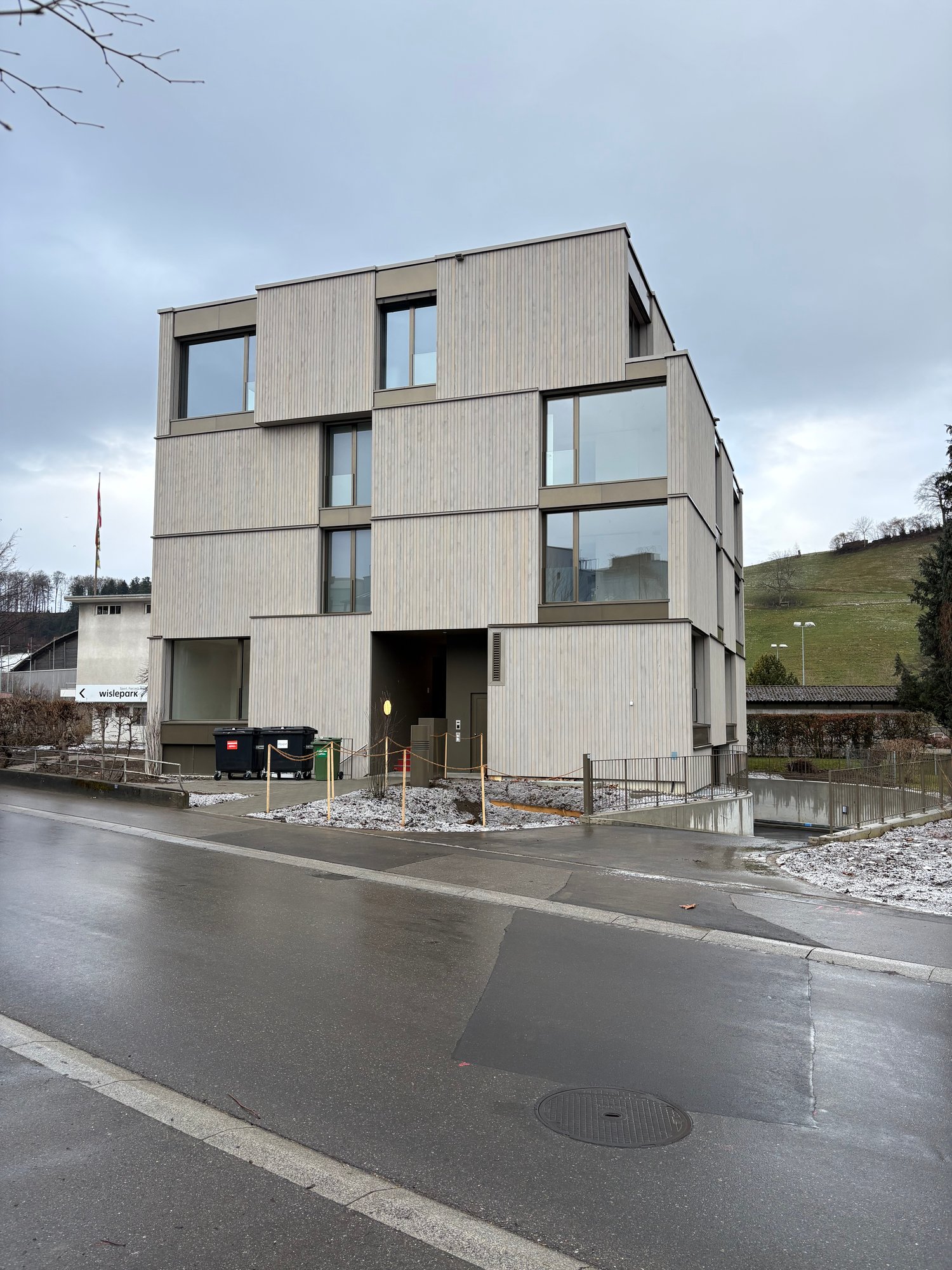 Multi-story residential building with glass windows, front entry, gray walls, fenced entrance, multiple trash bins, street light in the distance, and hilly area beyond