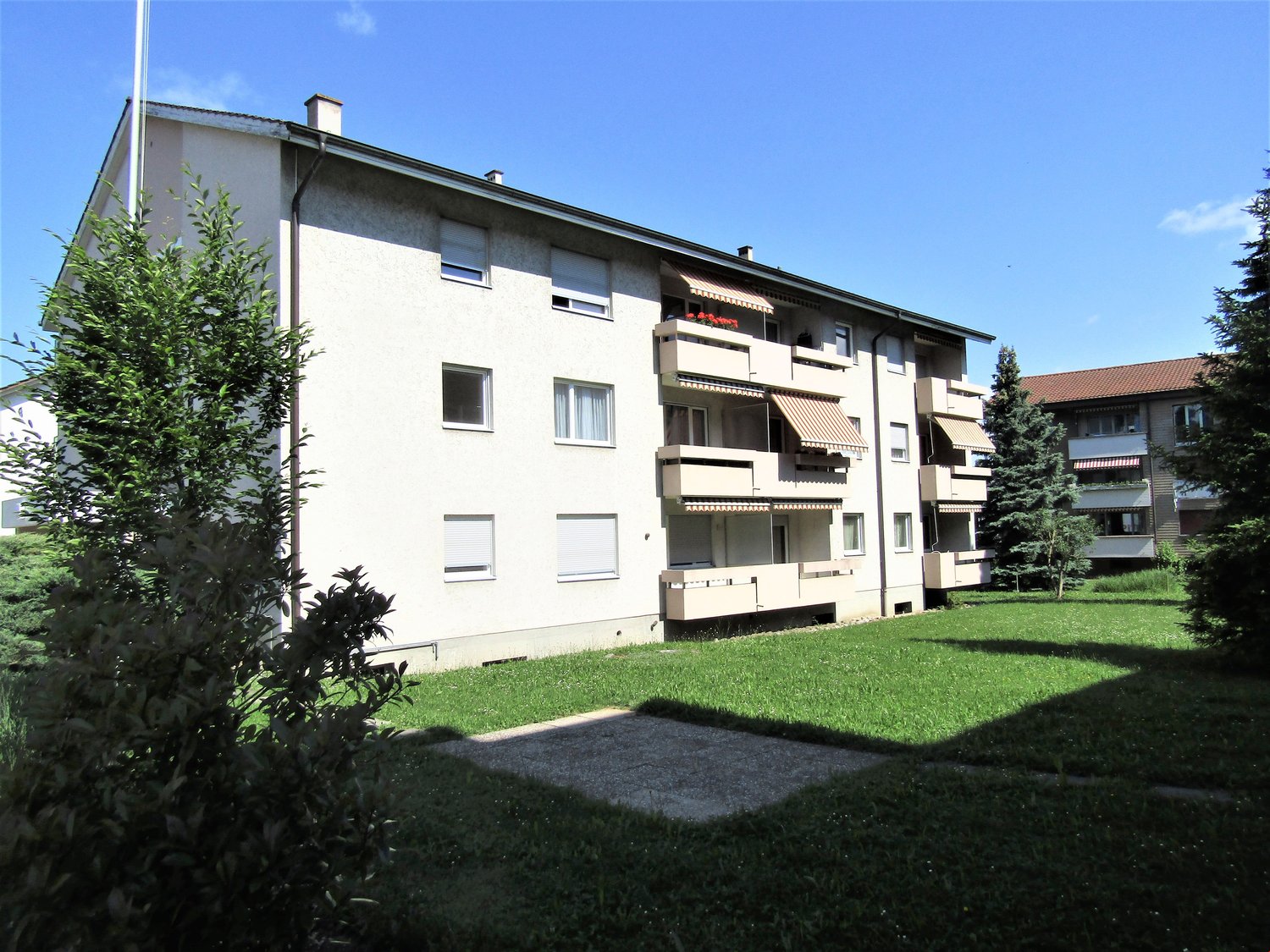 White multi-storey building, balconies with awnings, trees, and well-maintained lawn in front