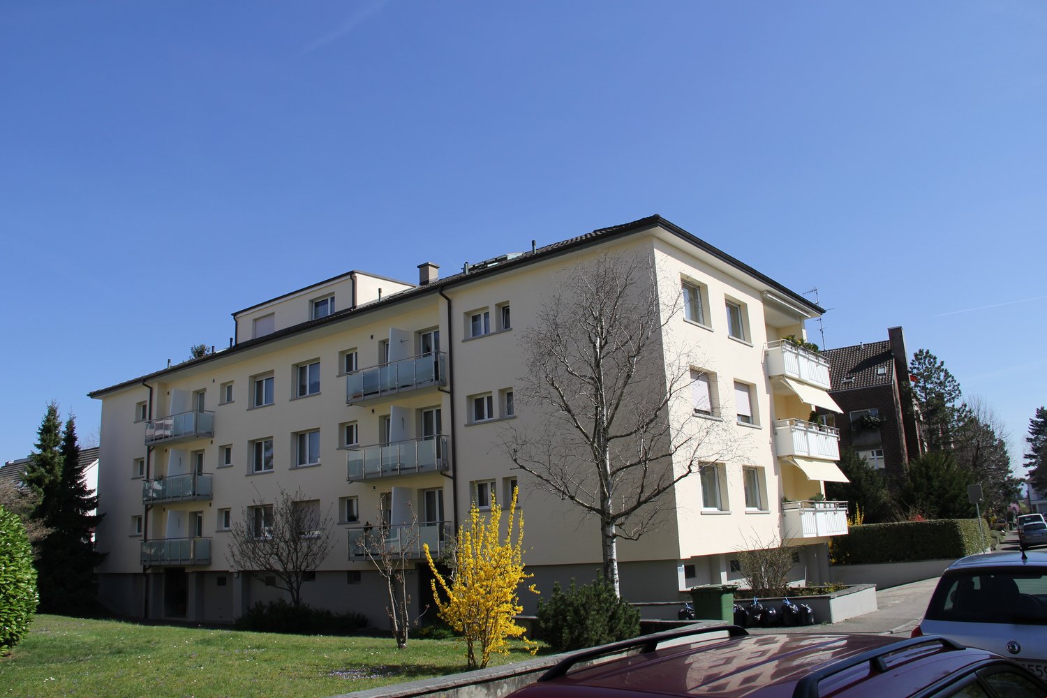 4 story apartment building, beige exterior, multiple balconies, windows, green bushes, trees, cars parked