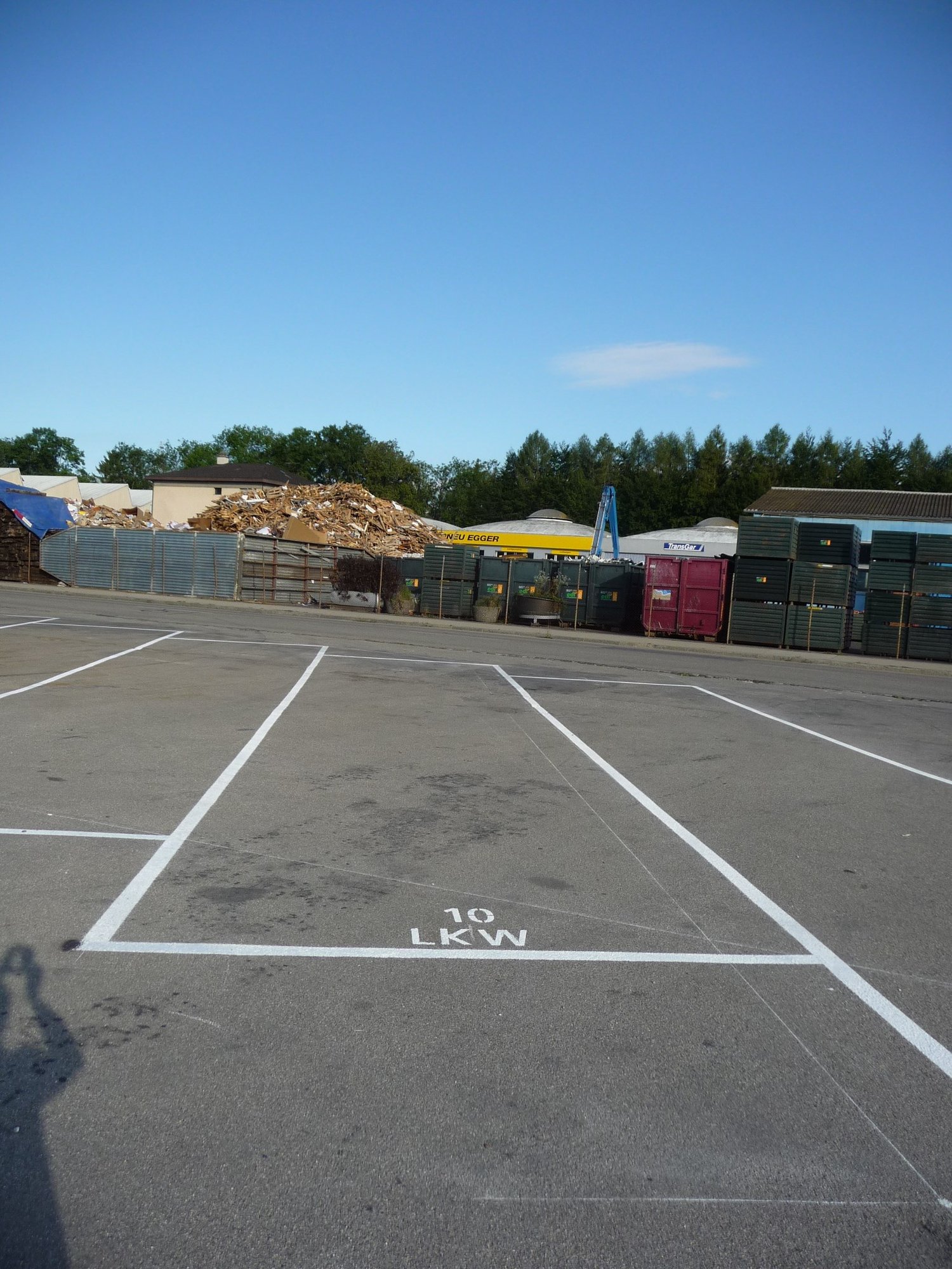 Paved parking lot with marked parking spaces, a large pile of wood debris, and various storage containers or dumpsters in the background.