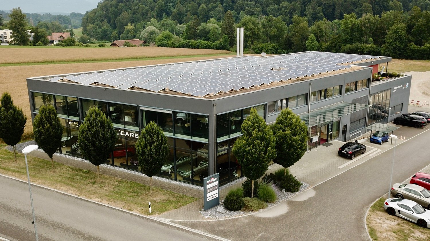 Commercial building with solar panels, multiple cars visible in windows, many cars parked outside, landscape with farm and trees