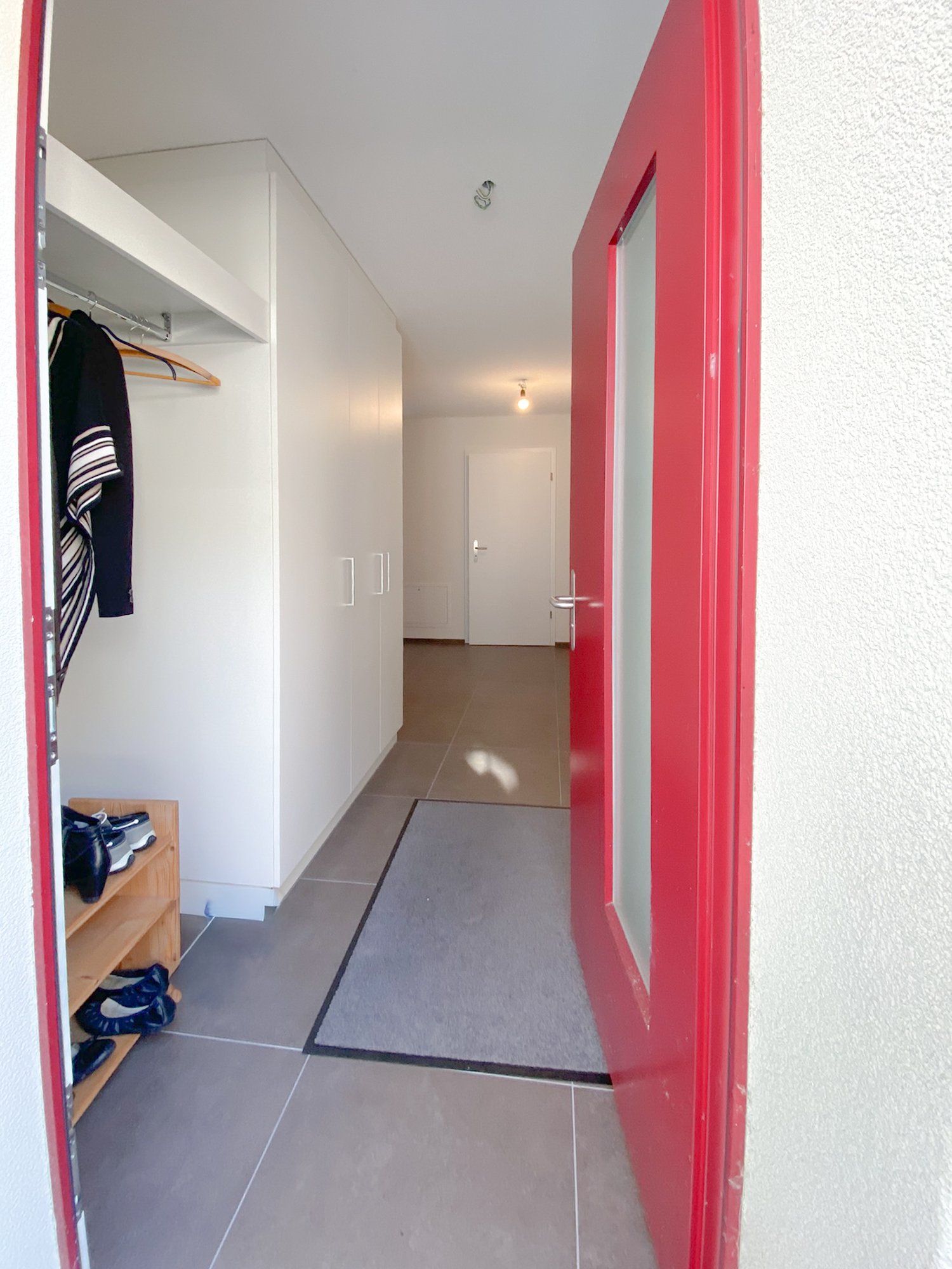The entryway of a building with red door, tiled floors, a small carpet, a closet with clothes and shoes visible, a wooden shelf with shoes, and a hallway leading to a white door. The hallway is illuminated by a light fixture on the ceiling. The entire int