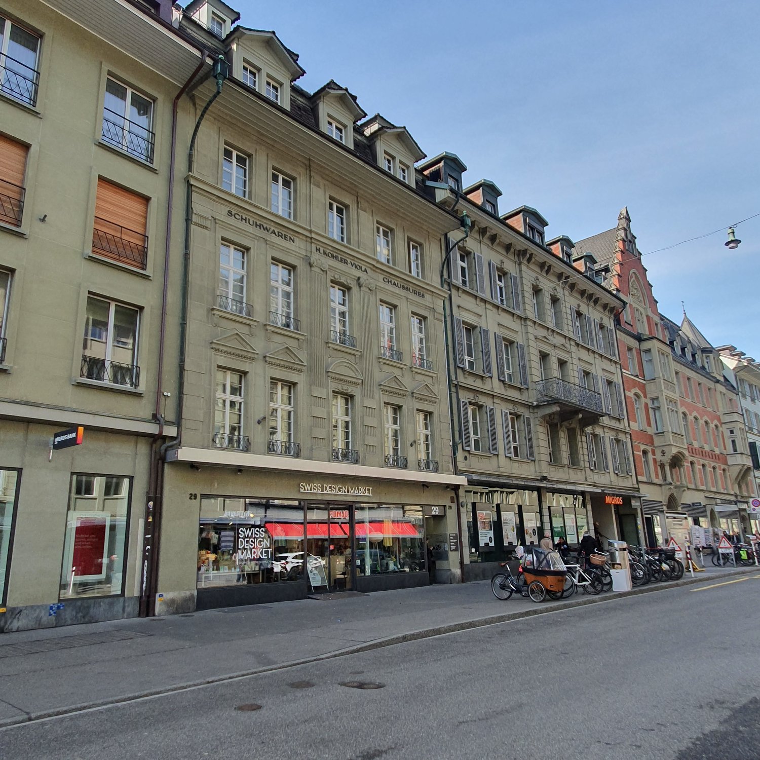 Multi-story apartment building on the city street, several shops on the ground floor with various storefronts, bicycles parked in front