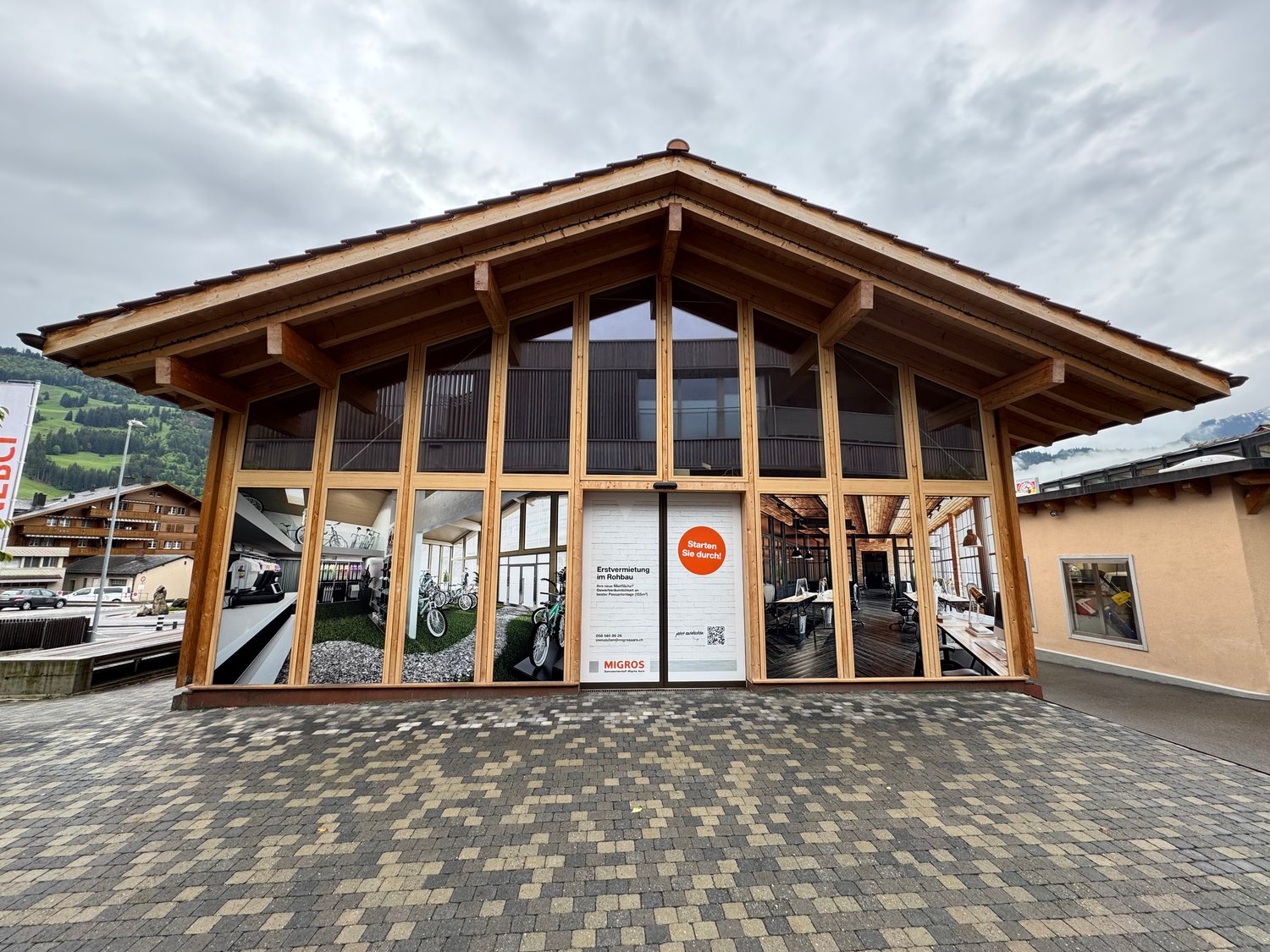 A large wooden building with a slanted roof and large windows. The building has a paved area in front of it, indicating a parking space. The building appears to be a commercial or public facility, with signage visible on the exterior.