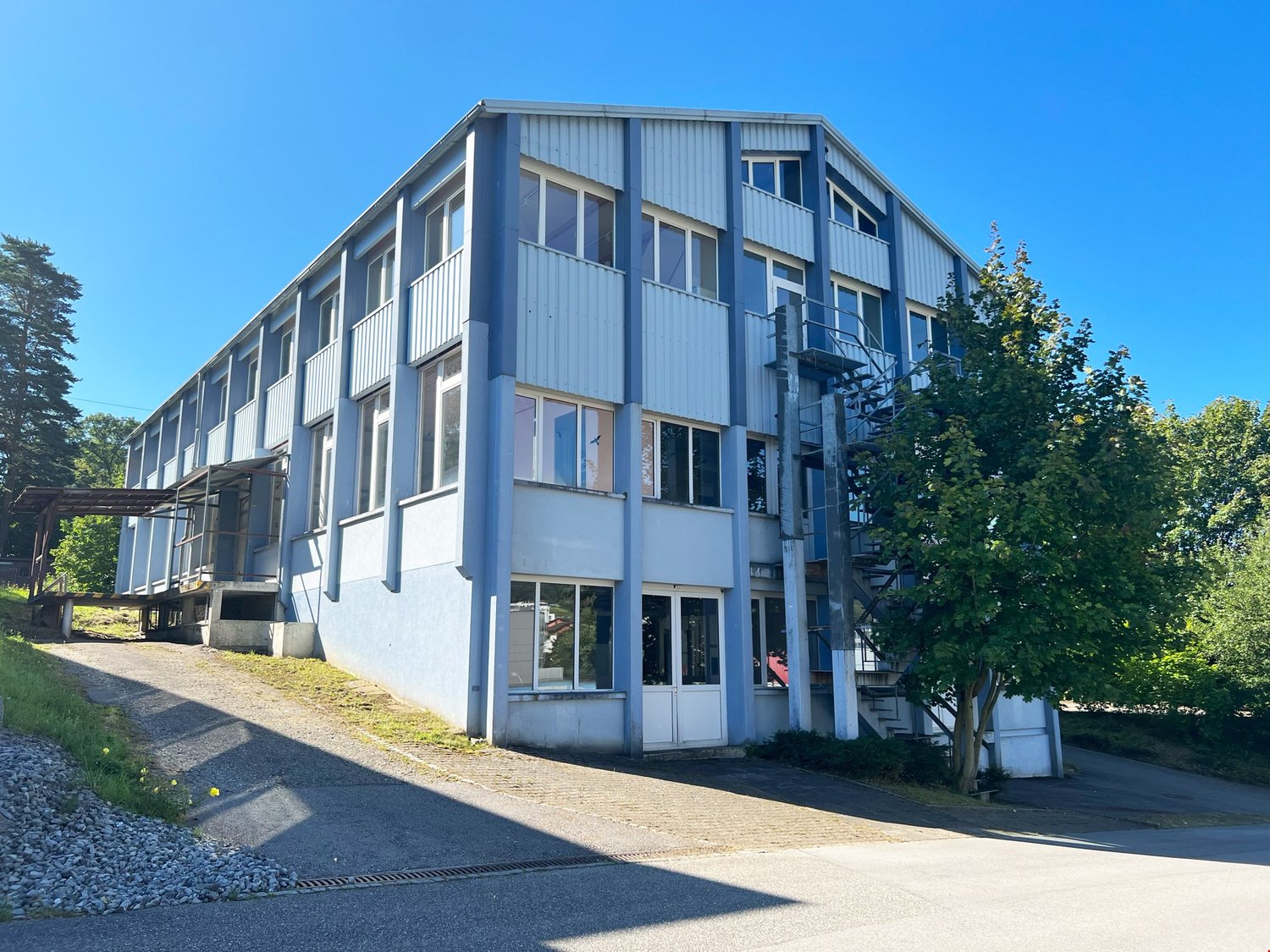 A multi-story commercial or industrial building with a gray metal exterior, large windows, and a fire escape on the side. The building is surrounded by trees and has a paved walkway leading up to the entrance.