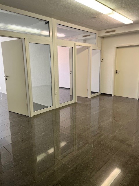The image shows an empty office space with a tiled floor and glass-paneled doors. The room has a clean, modern appearance with neutral-colored walls and overhead lighting.