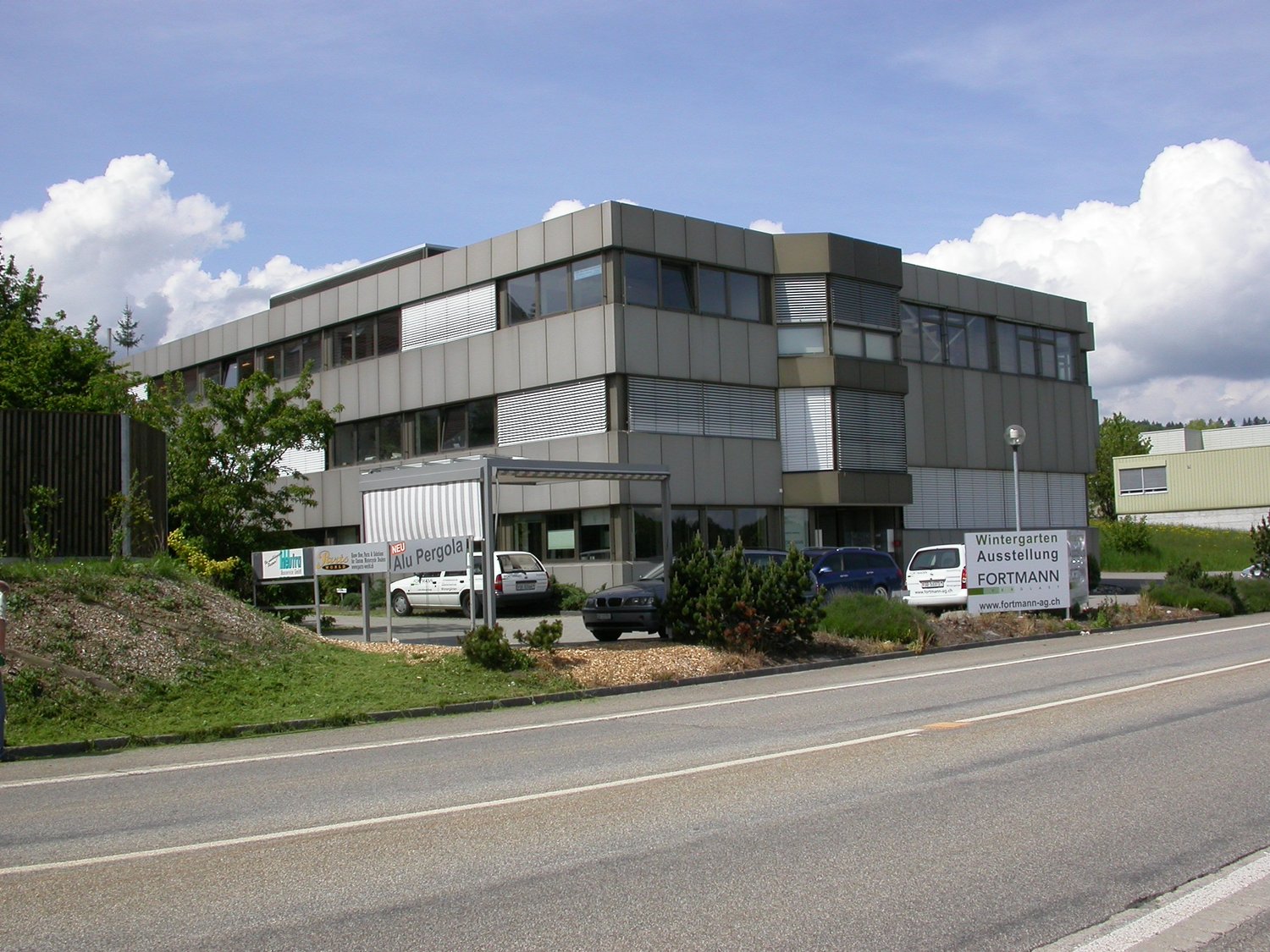 Commercial building, grey facade, large windows, cars parked in front, greenery around