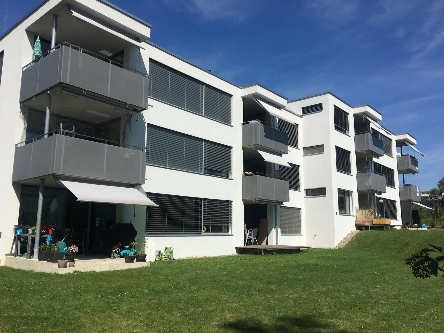 Modern white apartment building with multiple balconies, well-maintained garden, and outdoor seating area.