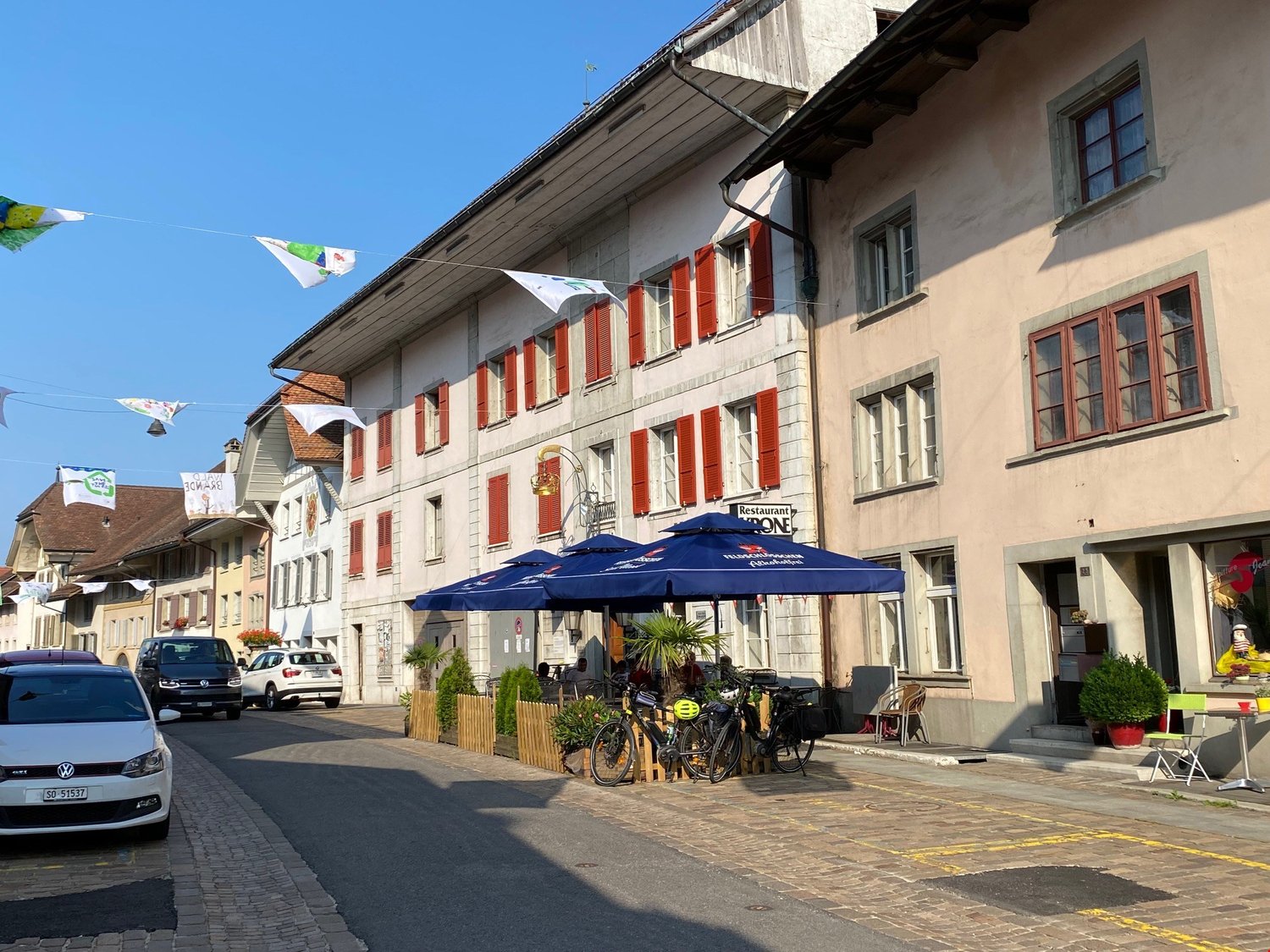 A street with an outdoor cafe area, bicycles, and cars parked. The buildings have red shutters and a decorative sign in front.