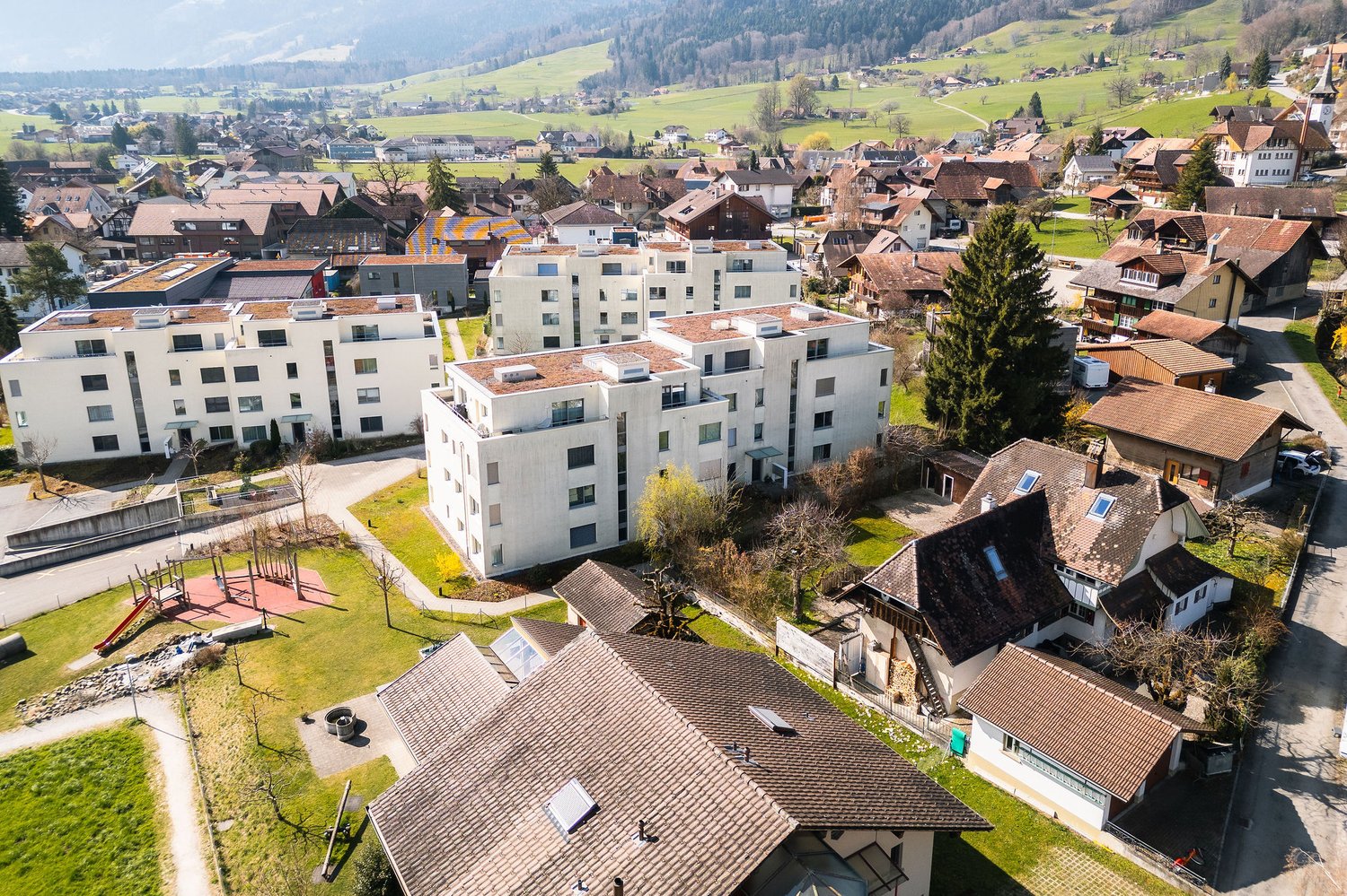 Aerial view of a residential area with multiple buildings, some with brown roofs and others with white facades, surrounded by green fields and trees.