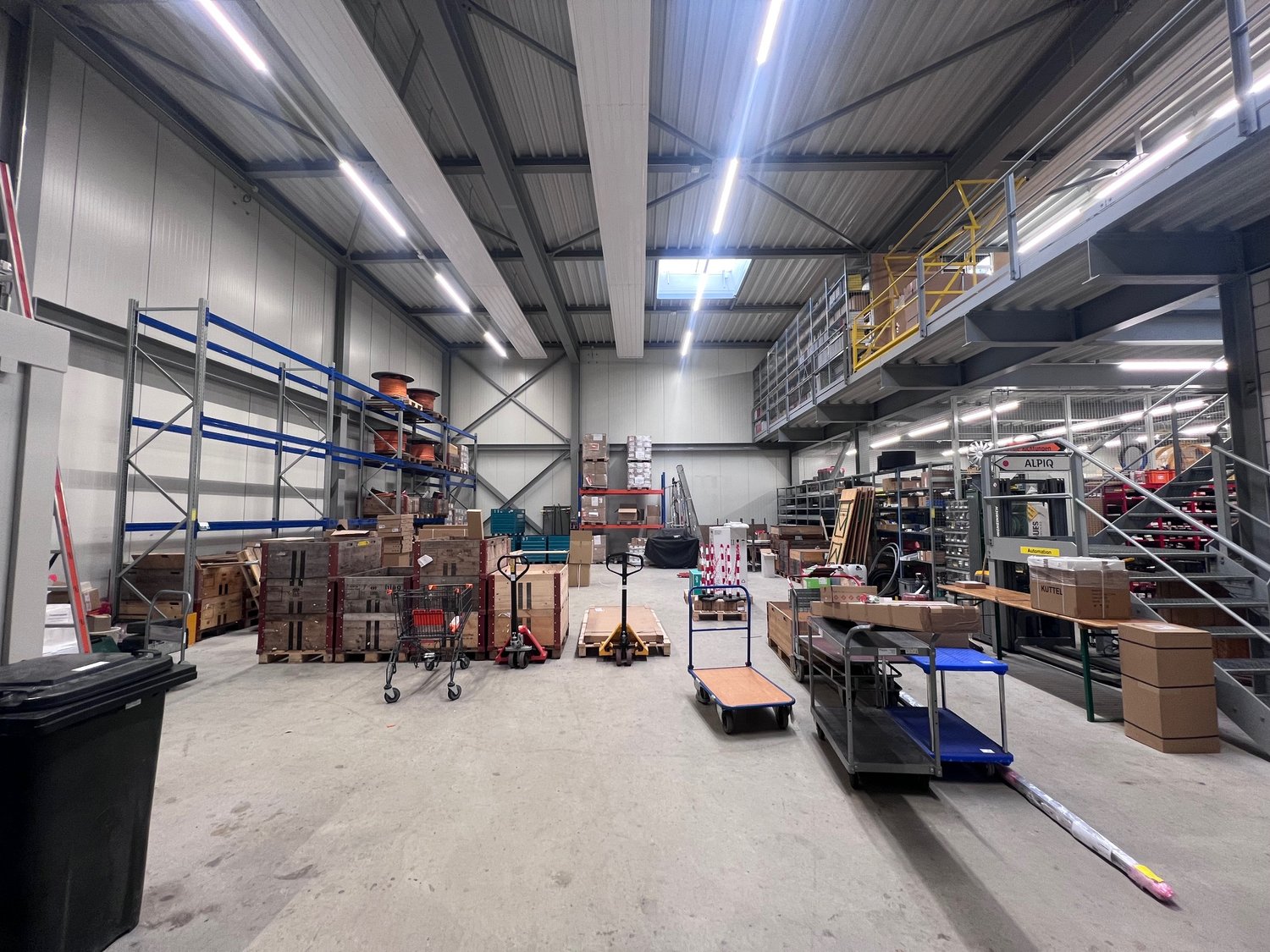 Spacious warehouse with high ceilings, multiple shelves and storage racks, various boxes and materials, trolleys and pallets, stairs, and metal beams.