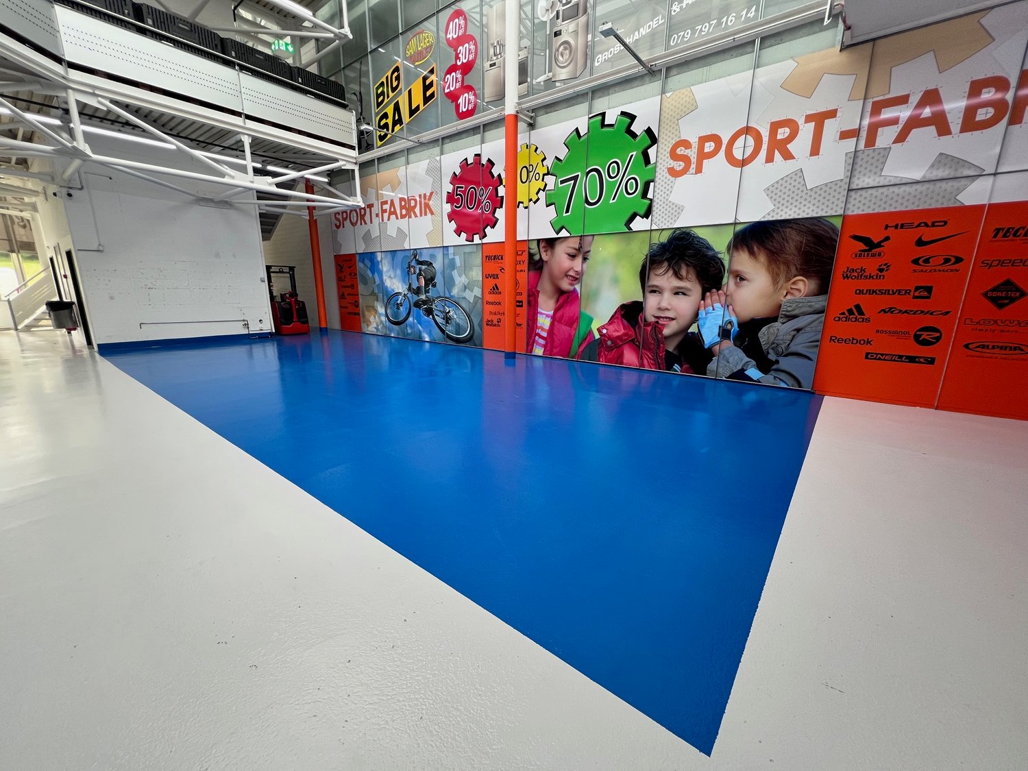Large indoor retail space with blue flooring, various sports and outdoor equipment brands displayed on the walls, and signs advertising sales and discounts.