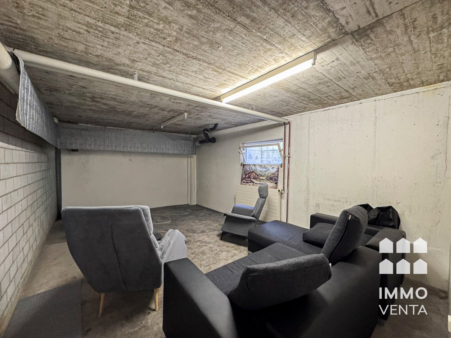 Empty room with concrete ceiling, white walls, gray floor, two couches, two armchairs, and a window with a picture