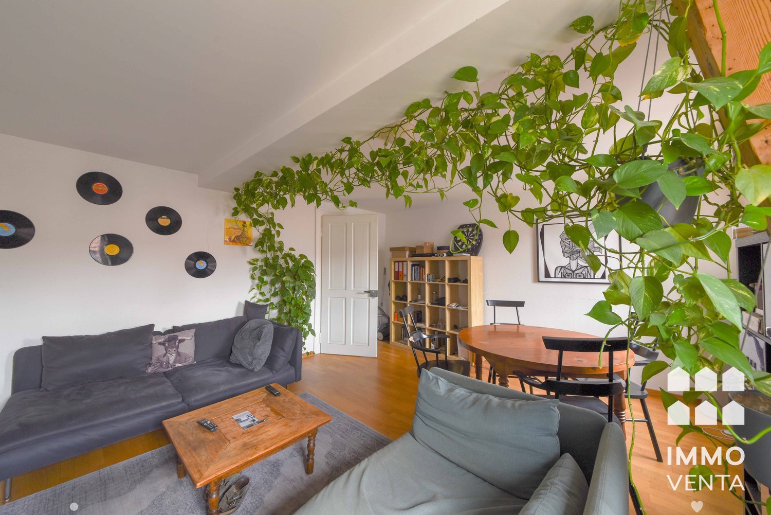 Living room with couch, coffee table, dining table, chairs, bookshelf, plants, vinyl records on wall