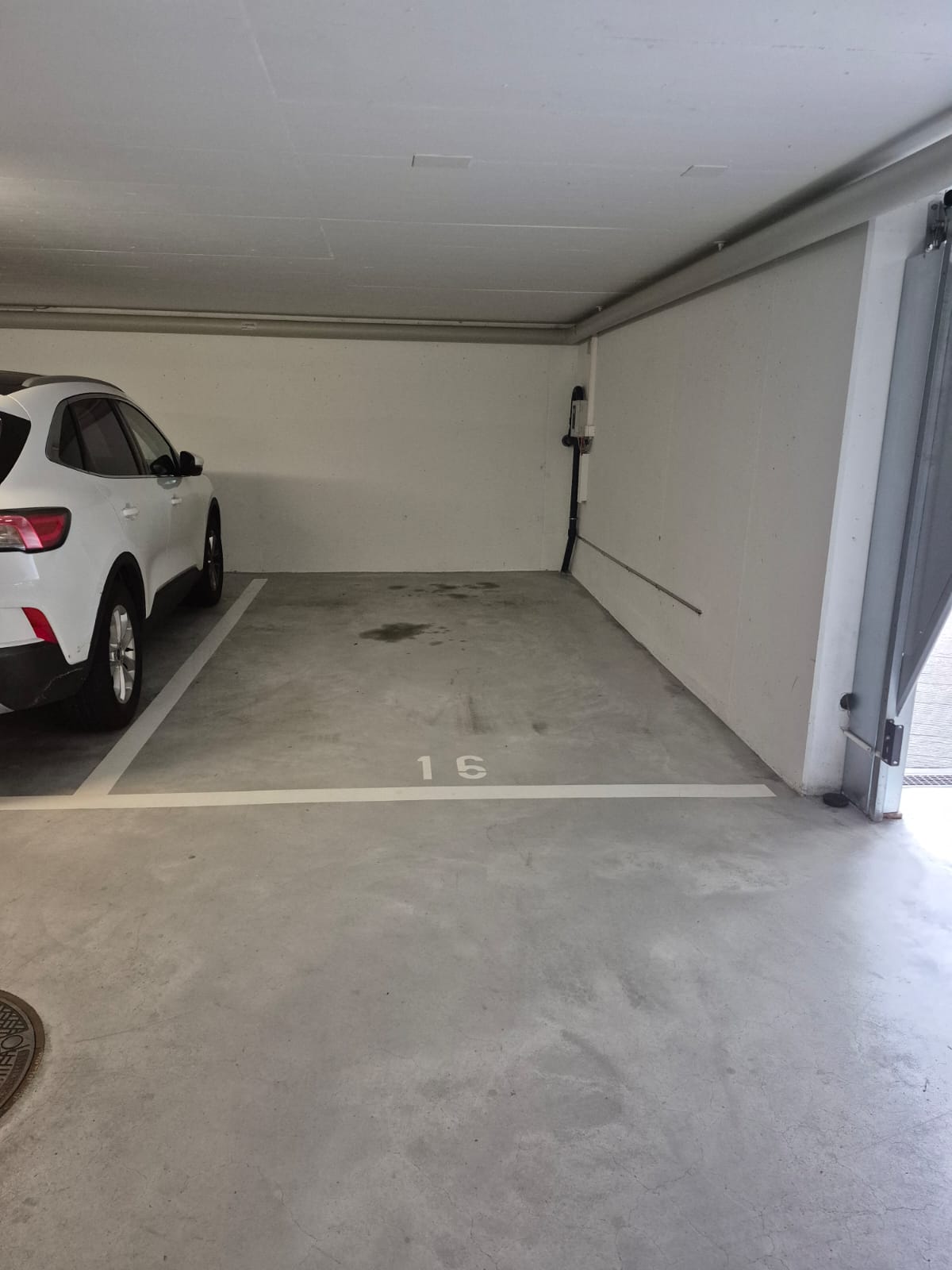The image shows a garage space with a white SUV parked inside. The floor is concrete and there is a number '15' painted on the floor, indicating this is parking spot number 15. The walls are plain white and there are some utility pipes or wires visible on