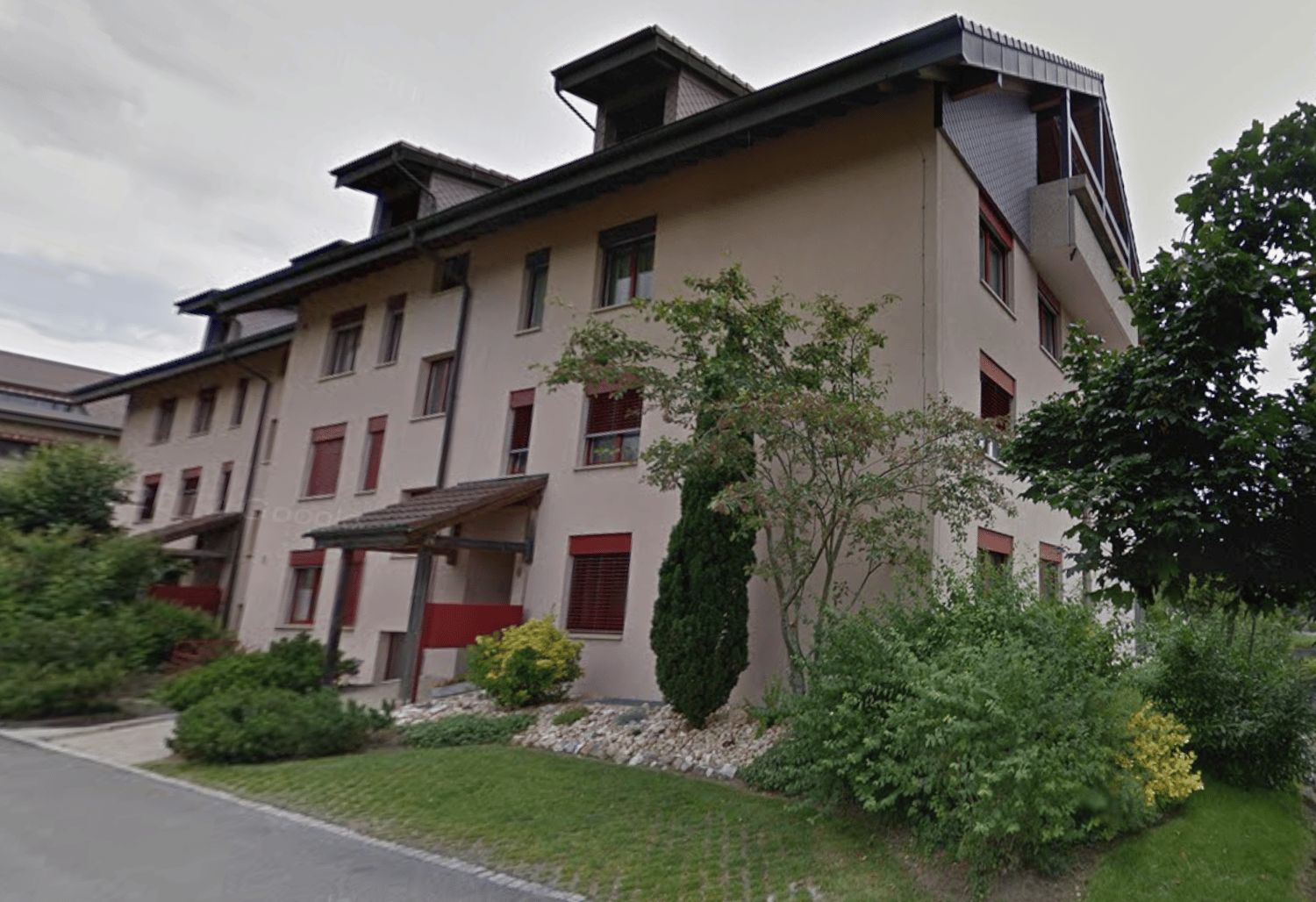 4-story beige building with red window frames, small bushes and trees in front
