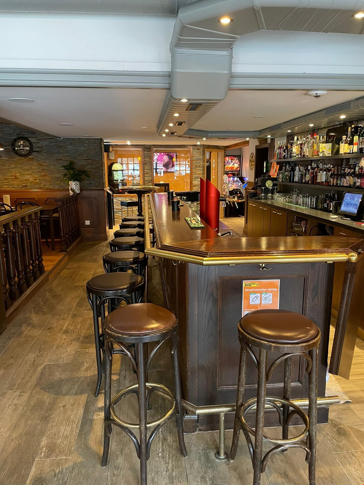 bar counter, stools, wooden flooring, wooden bar, bartop, shelves with alcohol bottles, gaming machines, plants