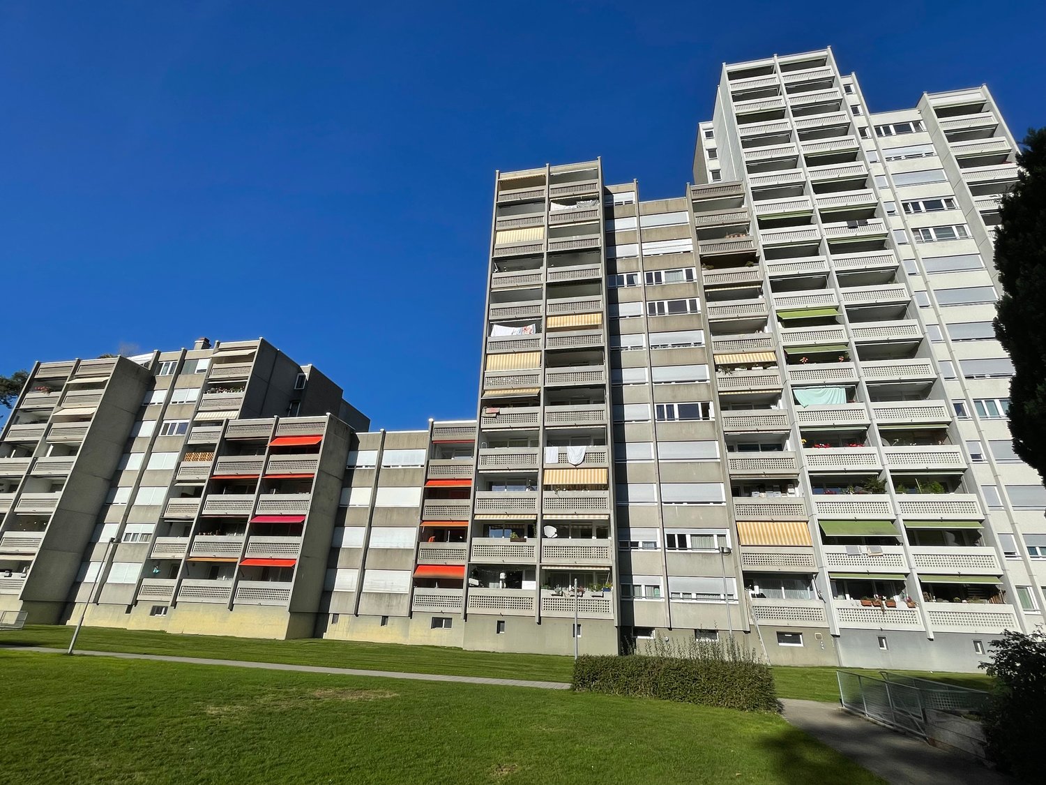 high rise apartment building, multiple floors, concrete exterior, red awnings, balconies, windows, greenery in front