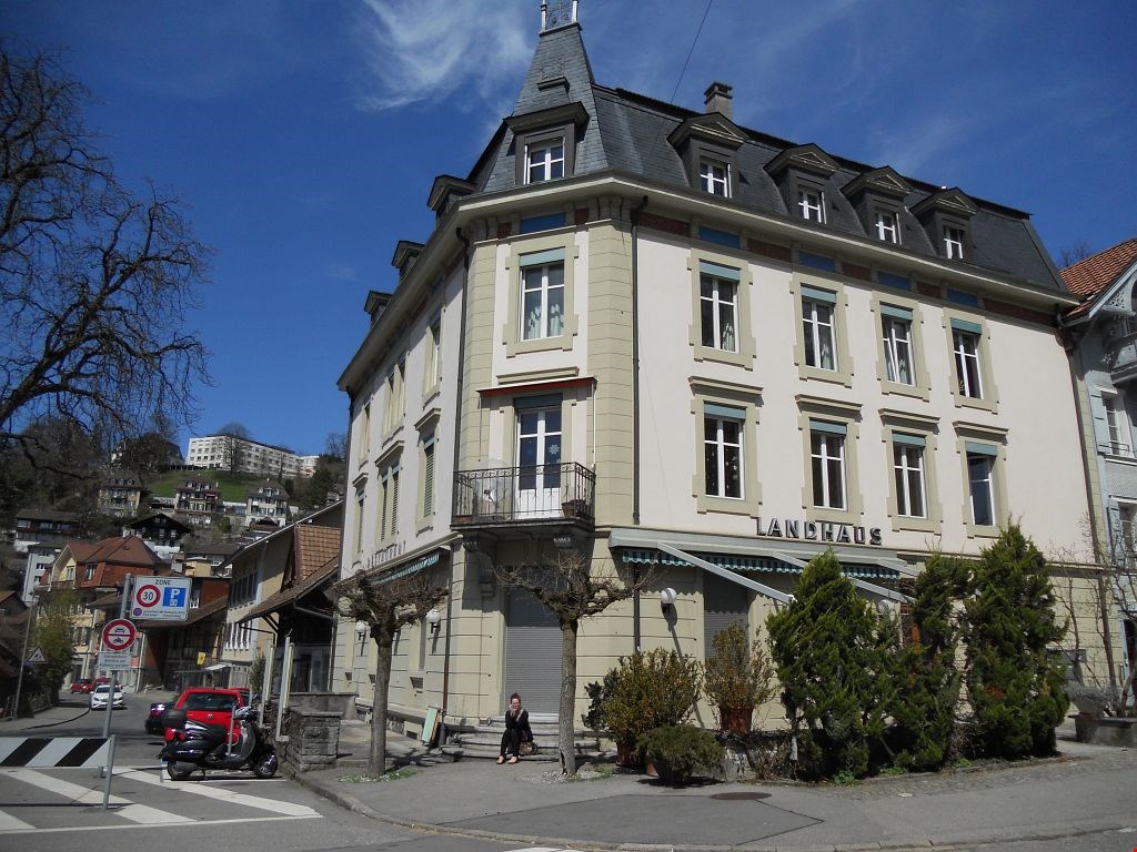 Corner building, 3 stories, white facade, multiple windows, beige trims, black roof, balcony with railings, awnings, and a name 'LANDHAUS' on the facade