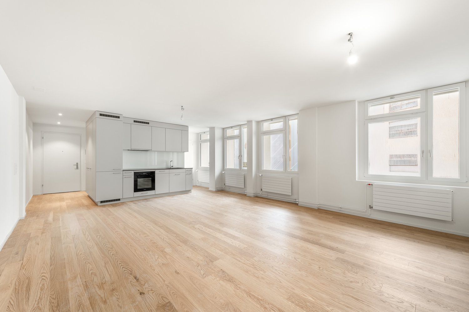 An empty kitchen with wooden floors, white walls, and large windows.